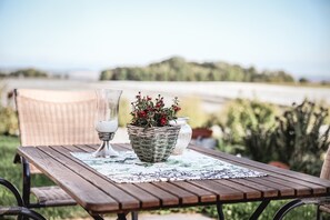 Ein Holztisch mit Untersetzer, ein Korbwagen mit Blumen, eine weiße Vase und ein Korbstuhl.