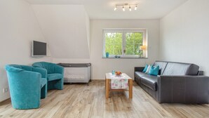Een woonkamer met een bank, twee fauteuils, een kleine tafel met een bloemenonderzetter, een radiator, een aan de muur bevestigde televisie en een raam met uitzicht op bomen.
