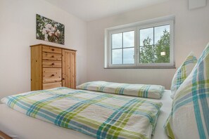 Une chambre avec deux lits, une commode en bois, une fenêtre donnant sur des arbres et un tableau floral accroché au mur.