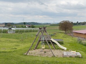 Un paysage rural avec une balançoire en bois, un petit toboggan et une zone délimitée par une clôture, avec des champs agricoles en arrière-plan.