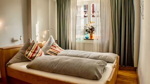 Un lit double avec une literie à rayures, une table de chevet avec une lampe, une fenêtre avec des rideaux et une vue sur les bâtiments à l’extérieur.