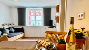 Un salon chaleureux avec un canapé, une table à manger agrémentée d’une bouteille de vin et d’un verre, un fauteuil et une fenêtre avec des volets.