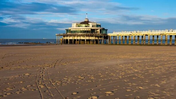 A beach with a pier extending into the sea, featuring a building with a dome and a flag on top.