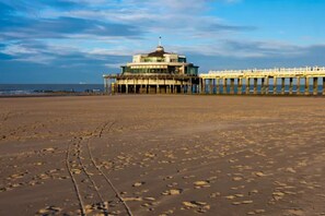 Une plage avec une jetée qui s’avance dans la mer, sur laquelle se trouve un bâtiment surmonté d’un dôme et d’un drapeau.
