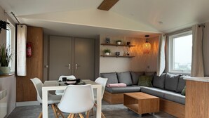 A modern living room with a dining area, a sofa, and a coffee table. There is a window with curtains, a shelf with plants, and a hanging light fixture.