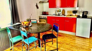 A compact kitchen with red cabinets, a small stove, and a refrigerator. There is a dining table with chairs and a window with curtains.