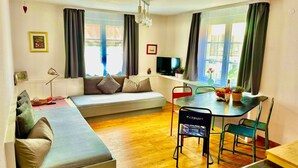 A living room with a sofa, a dining table with chairs, a TV, and large windows with curtains.