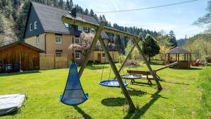 Een houten schommelset, een trampolaan en een houten speeltuintje in een achtertuin, met een huis op de achtergrond.