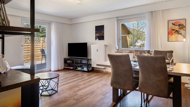 Een moderne woonkamer met houten vloer, een eettafel met stoelen, een flatscreen televisie en een boekenkast.