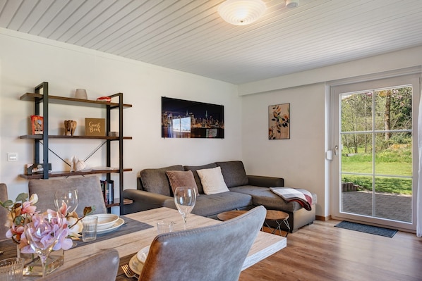 Una sala de estar moderna con un sofá, una mesa de comedor con sillas, una estantería y un amplio ventanal con vistas al verdor.