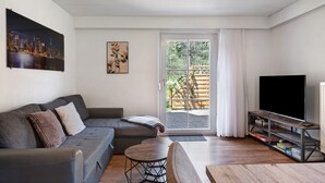 Een moderne woonkamer met een grijze bank, een klein rond tafeltje, een tv op een houten dressoir en een schuifpui naar een buitenruimte.