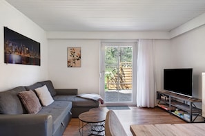 A modern living room with a grey sofa, a small round table, a TV on a wooden stand, and a sliding glass door leading to an outdoor area.