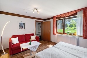 Ett rum med en röd soffa, ett litet bord med en vas, en säng, ett fönster med gardiner och ett element.