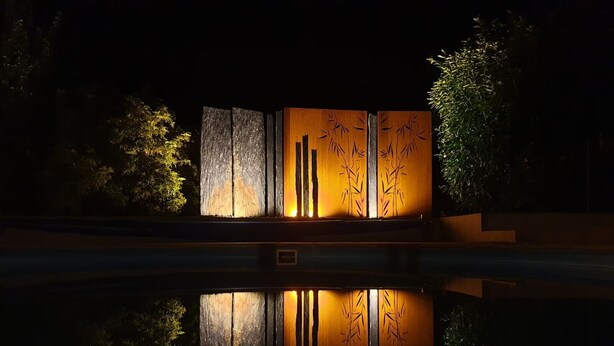 Ein glitzernder Pool mit einer Holzkonstruktion aus vertikalen Lamellen und Bambusmuster, umgeben von Vegetation.
