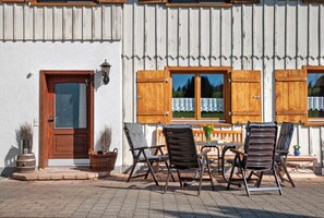 Un edificio con puerta de madera, ventana con cortina y asientos exteriores con sillas y una mesa.