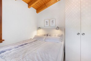 Une chambre avec un plafond en bois, un lit avec une literie blanche, deux lampes fixées au mur et une armoire.