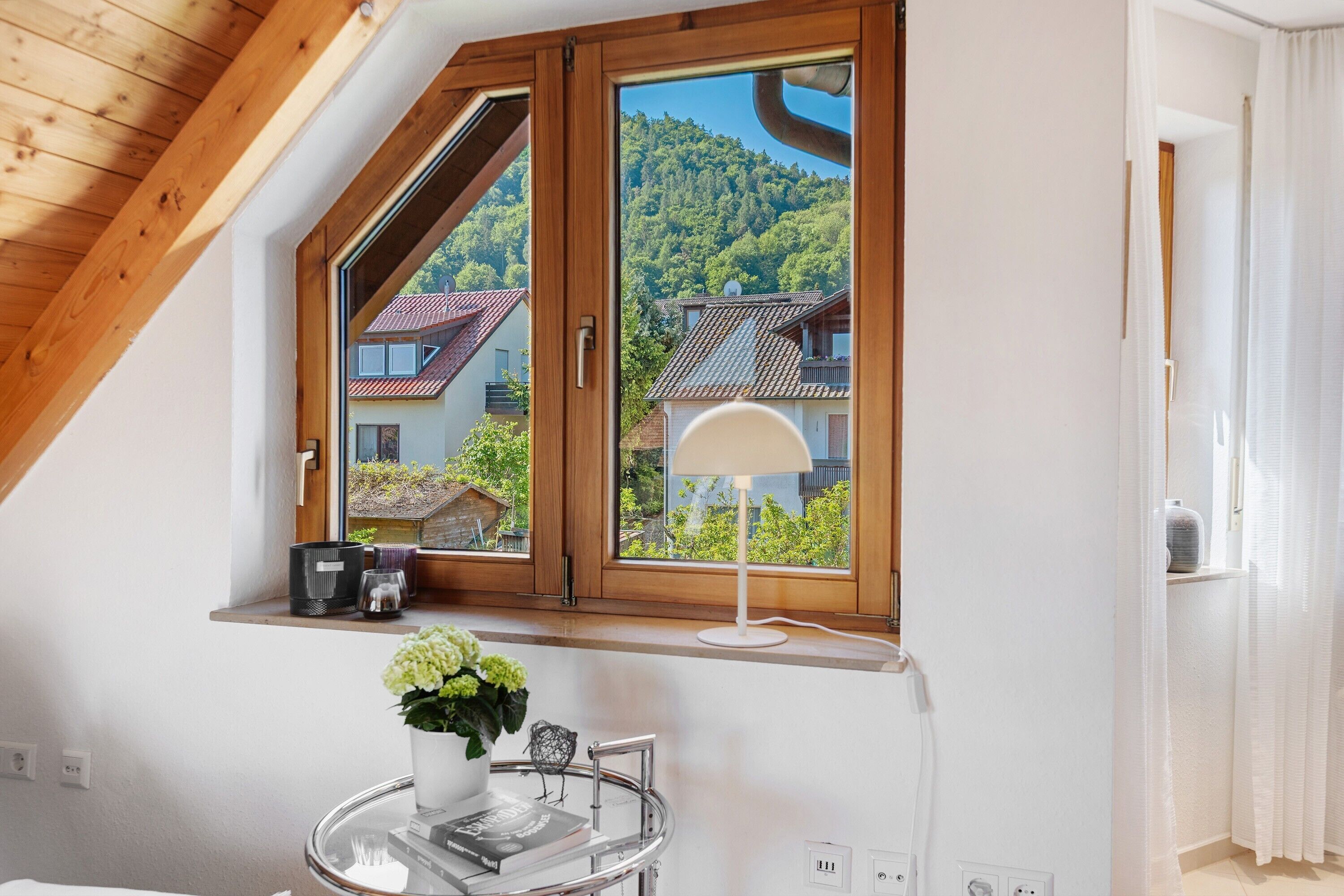 Een kamer met een schuine kap, een raam met uitzicht op huizen en groen, een tafel met een lamp en een vaas met bloemen.
