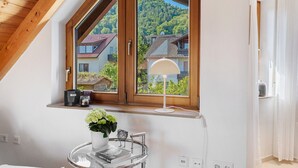 A room with a sloped ceiling, a window with a view of houses and greenery, a table with a lamp, and a vase with flowers.