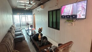 A hotel room with a long sofa, a small table with a vase, a chair, a ceiling fan, and a TV mounted on the wall. - Ganga Godawari Guest House (Varanasi)