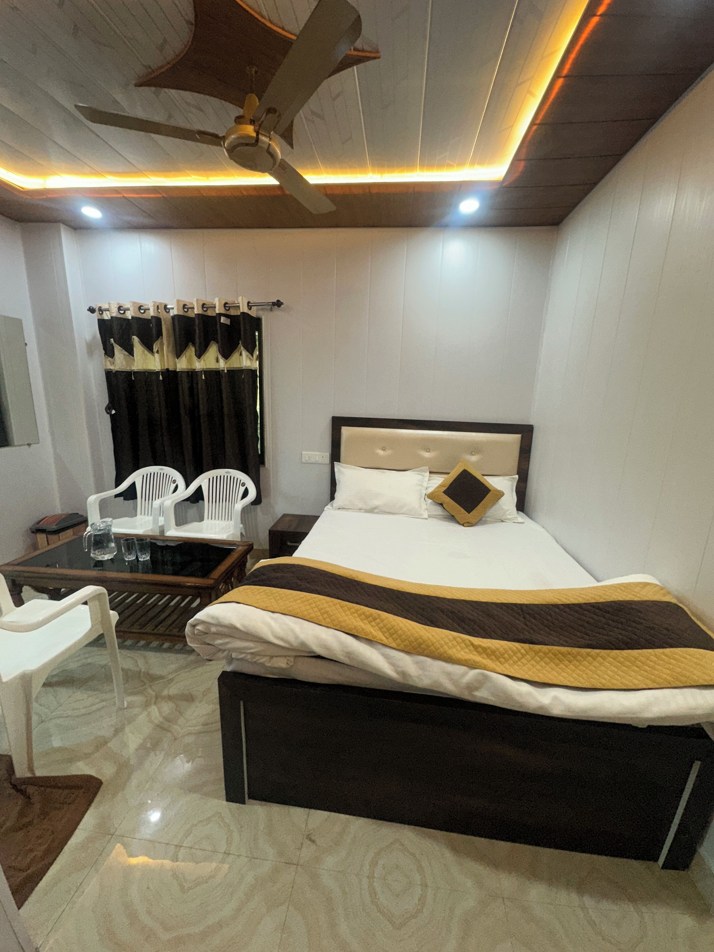 Una habitación de hotel con una cama, una mesa con sillas, un ventilador de techo y una ventana con cortinas.