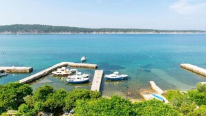 A marina with several boats docked at wooden piers, surrounded by lush greenery and a clear blue sea. - Studio in Rab near Sandy Beach (Rab)