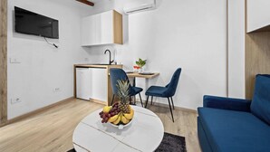 Una sala de estar moderna con un sofá azul, una mesa de centro blanca con frutas, un televisor montado en la pared y una zona de cocina con lavavajillas y una planta.