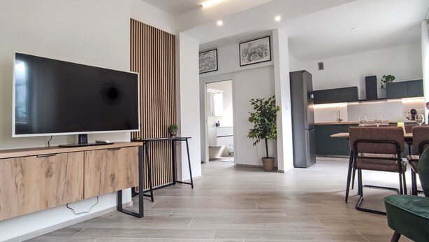 Ein modernes Wohnzimmer mit Flachbildfernseher, Wandpaneelen aus Holz und einem Essbereich mit Stühlen und Tisch.