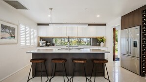 A modern kitchen with a central island, bar stools, a built-in wine cooler, and a view of greenery outside. - Beautiful ocean views, beaches and bushwalks (MACMASTERS BEACH)