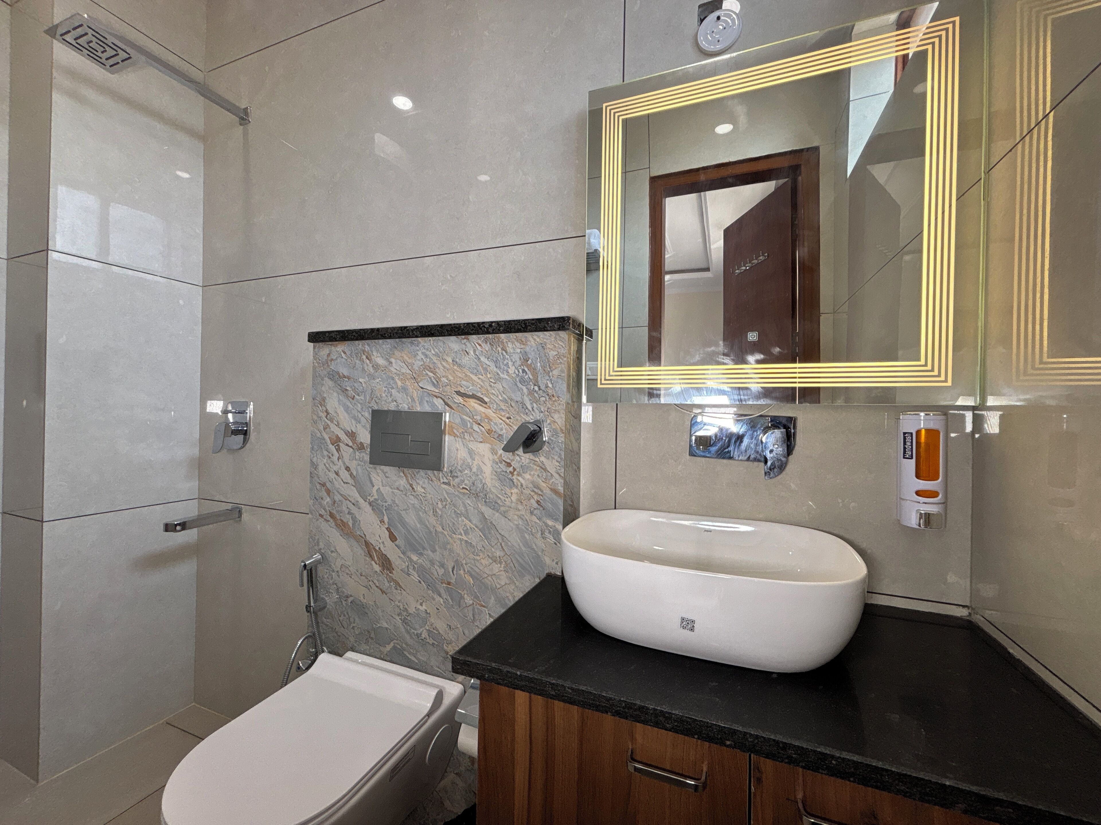 A modern bathroom with a marble wall, a large mirror, a white sink, and a toilet.