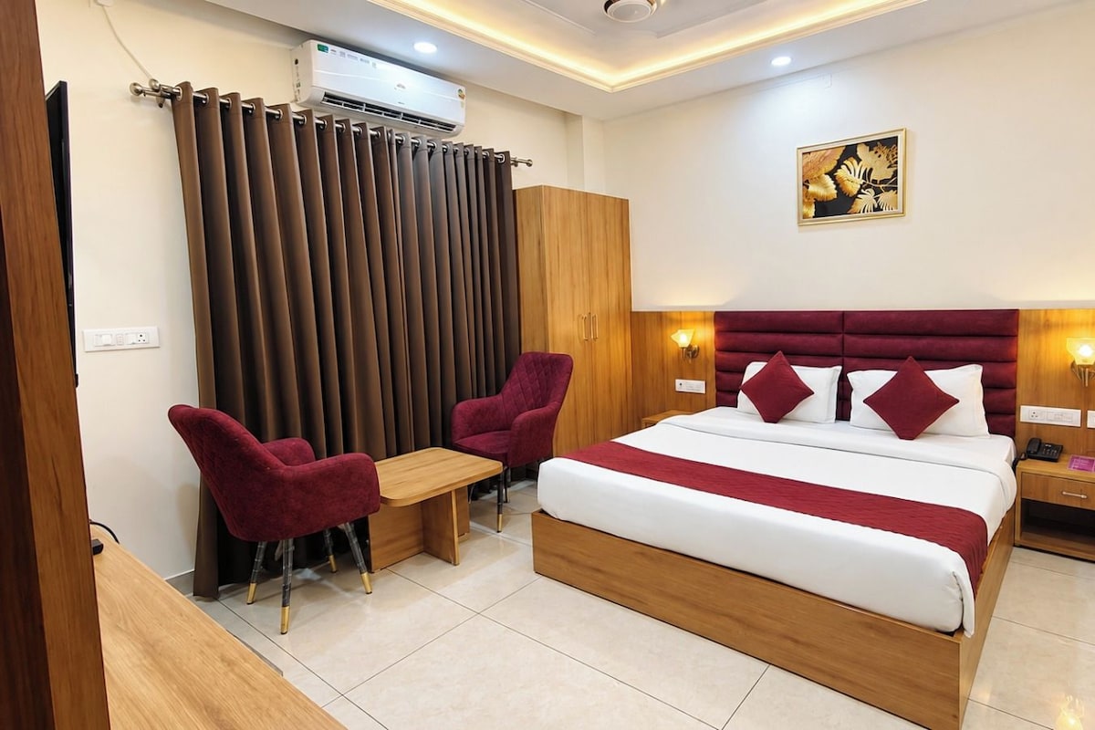 A hotel room with a bed, two red chairs, a wooden desk, a nightstand, and a wall-mounted air conditioner.