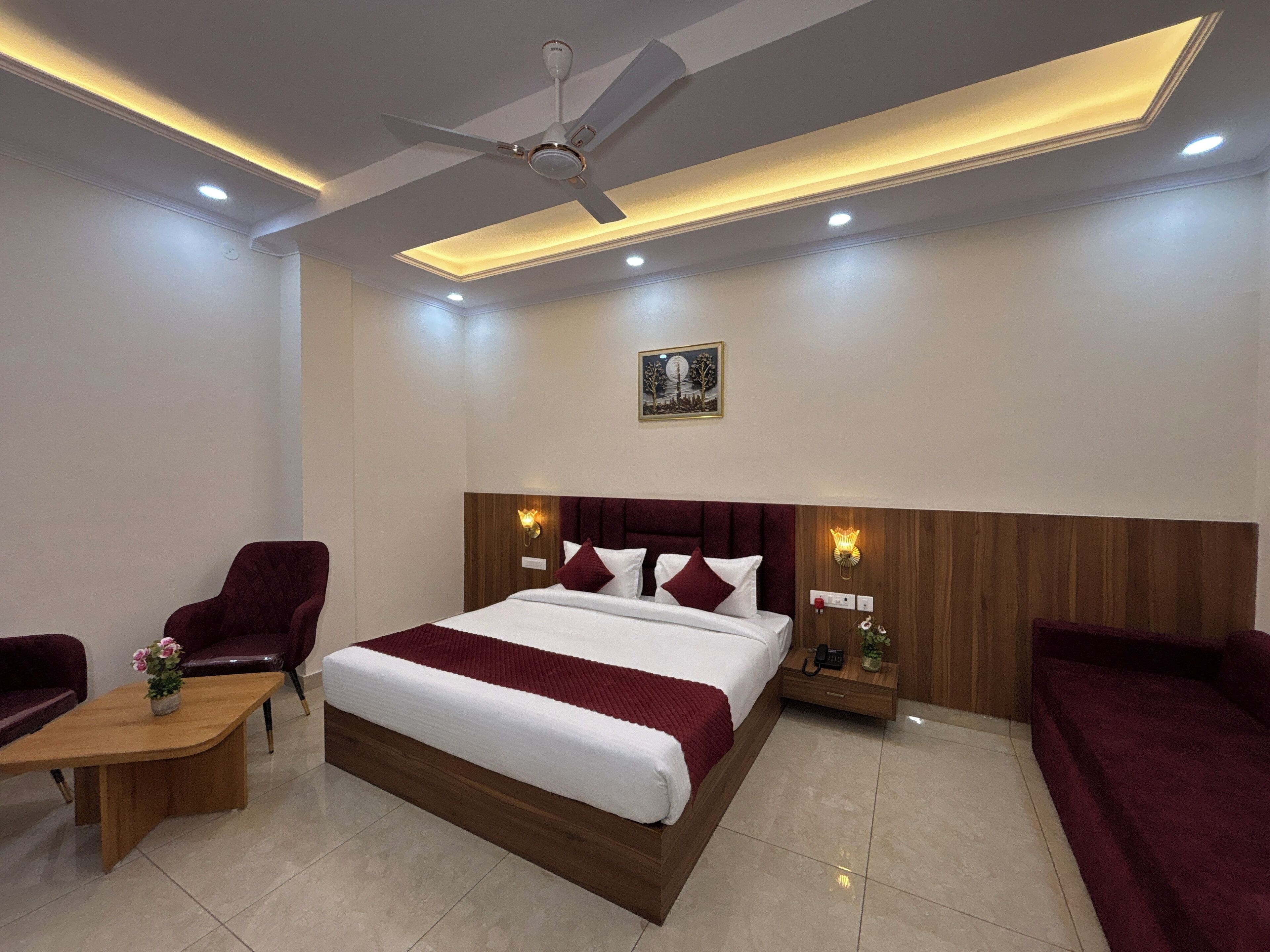 A hotel room with a large bed, two armchairs, a small table, a ceiling fan, recessed lighting, and a wall-mounted artwork.
