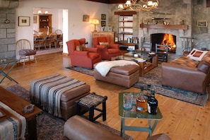 A cozy living room with a fireplace, leather sofas, a coffee table with bottles, and a dining area in the background.