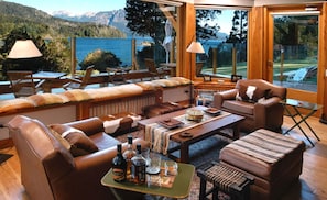 A spacious living room with leather furniture, a coffee table with bottles, and a view of a lake and mountains.