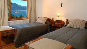 A bedroom with two beds, a wooden nightstand with a lamp, a window with a view of a lake and mountains, and a radiator.