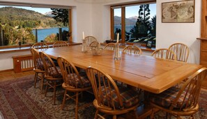 A dining room with a large wooden table, six wooden chairs with plaid cushions, and large windows offering a view of a lake and mountains. - Spectacular house by the Nahuel Huapi lake (San Carlos de Bariloche)