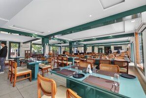 A spacious dining area with multiple tables and chairs, large windows, and a high ceiling with recessed lighting. - Corbett Selva Resort By DevStayz -Pet-Friendly Resort (Ramnagar)