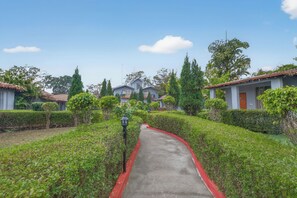 A pathway leading through a well-maintained garden with neatly trimmed hedges and a variety of trees. - Corbett Selva Resort By DevStayz -Pet-Friendly Resort (Ramnagar)