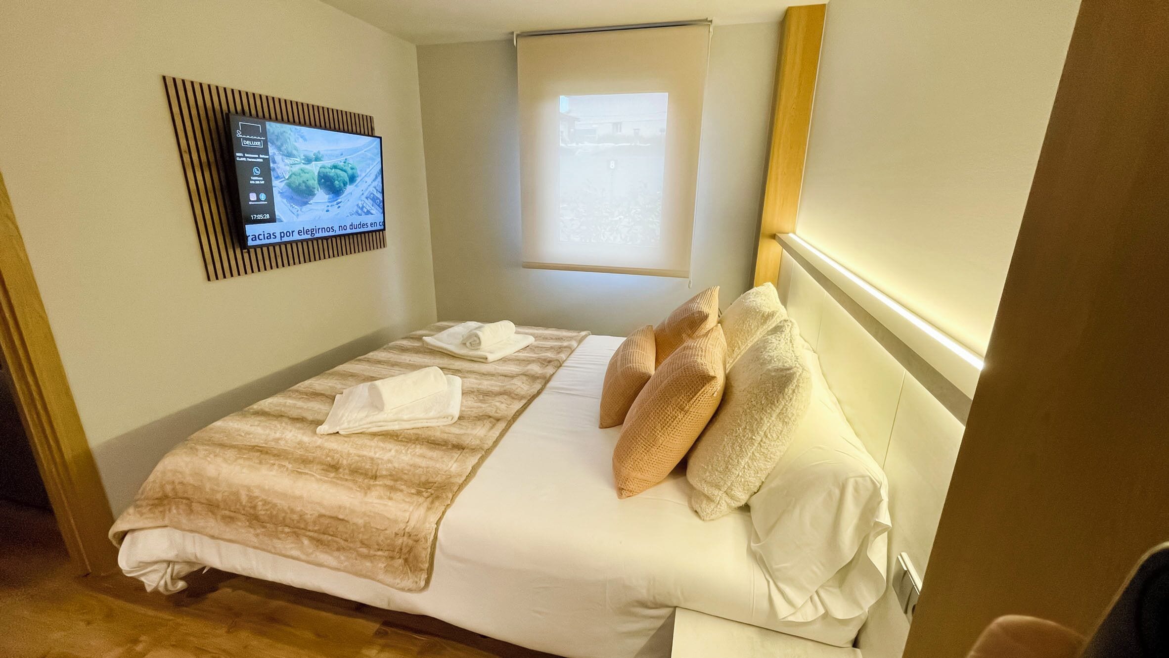 Habitación de hotel con cama, televisión montada en la pared y ventana con persianas.
