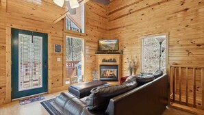 A log cabin interior with a fireplace, leather sofa, and a view of the outdoors. - KunTree Cabin by Beyond Expectations: 3BR Cabin w/ Hot Tub, Sauna & Pool Table (Sevierville)