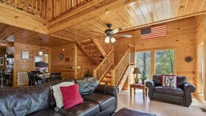 A wooden cabin interior with a leather sofa, a dining area, a staircase, and an American flag. - KunTree Cabin by Beyond Expectations: 3BR Cabin w/ Hot Tub, Sauna & Pool Table (Sevierville)