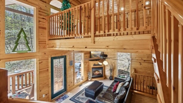 A wooden cabin interior with a loft bedroom, a living room with a fireplace, and large windows offering views of the surrounding forest. - KunTree Cabin by Beyond Expectations: 3BR Cabin w/ Hot Tub, Sauna & Pool Table (Sevierville)