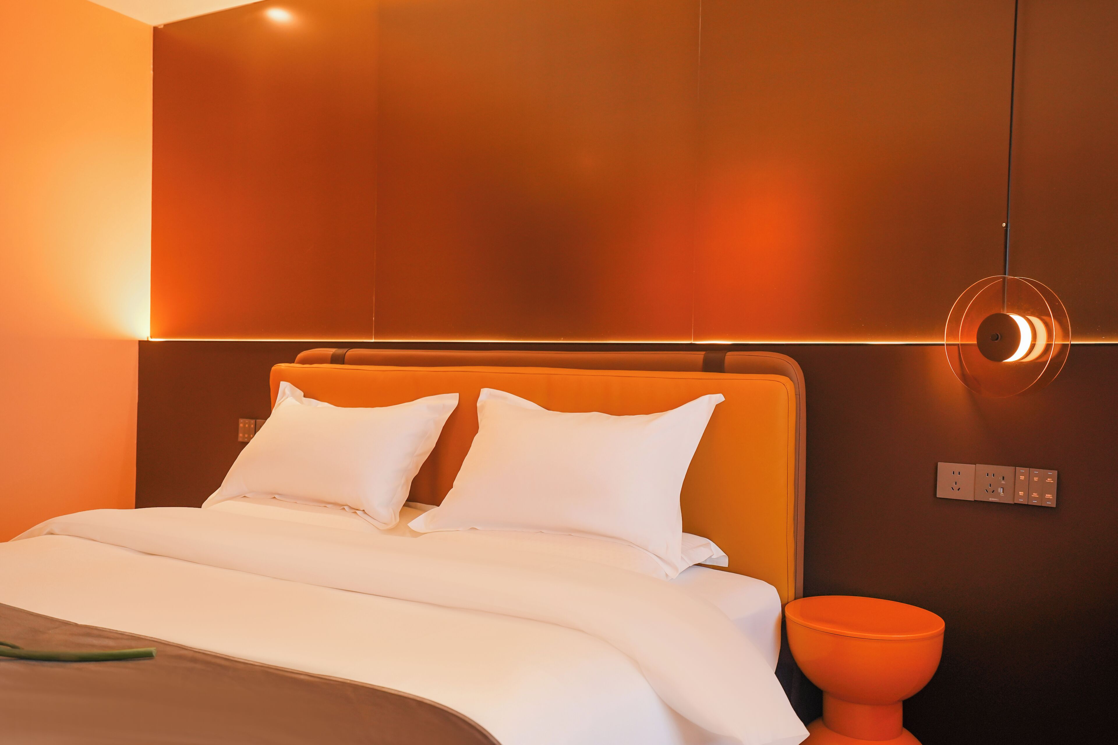 Una habitación de hotel moderna con una cama grande, ropa de cama blanca, una pared de acento naranja y una lámpara circular original.