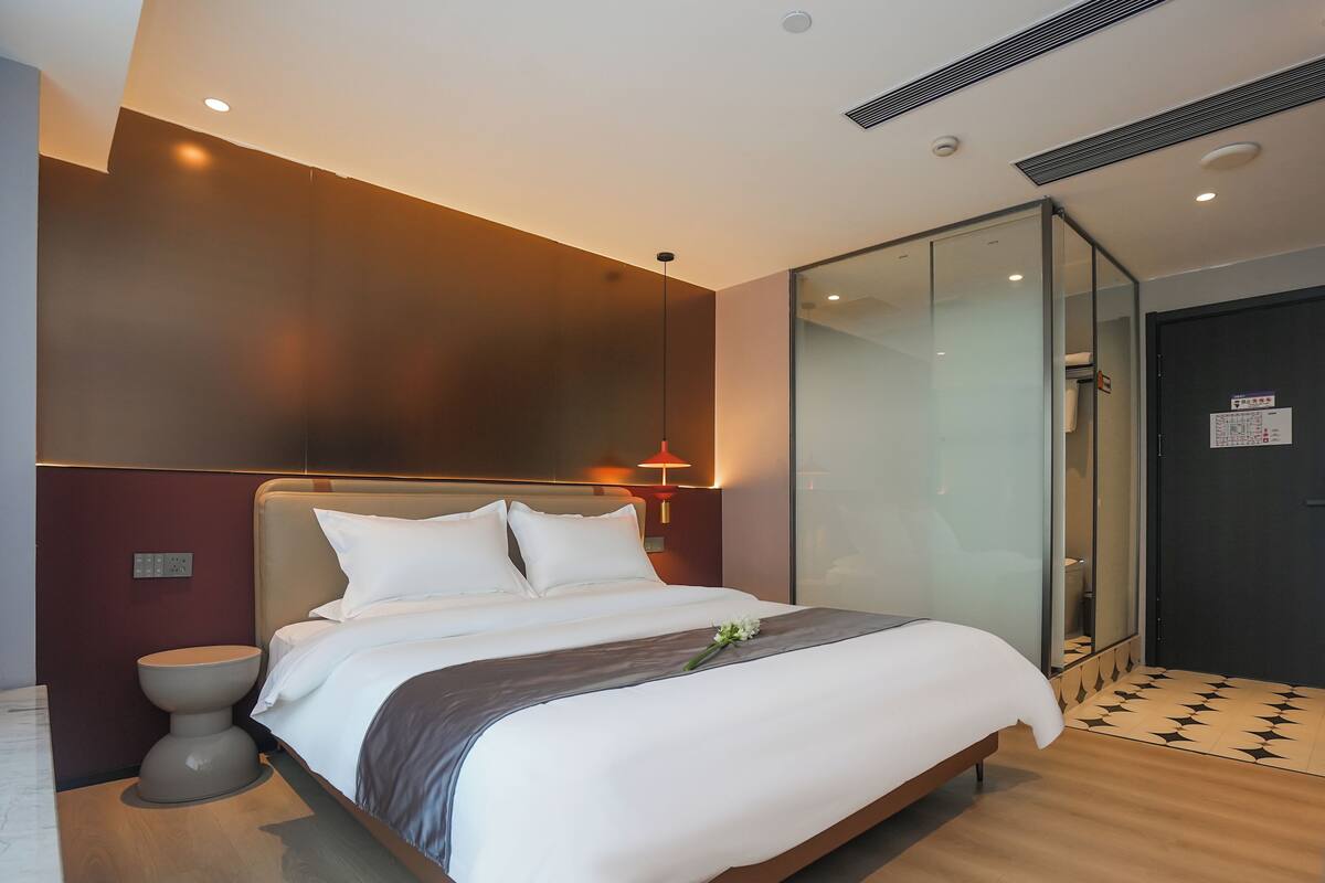 A modern hotel room with a large bed, a bedside table, a glass shower enclosure, and a dark door.