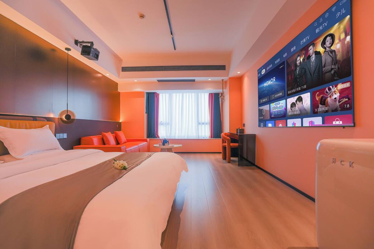 A modern hotel room with a large bed, a seating area with orange sofas, a TV wall displaying entertainment options, and a large window with curtains.