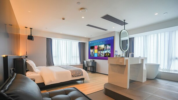Una habitación de hotel moderna con una cama grande, un sofá de cuero negro, un televisor montado en la pared y un amplio ventanal con cortinas.