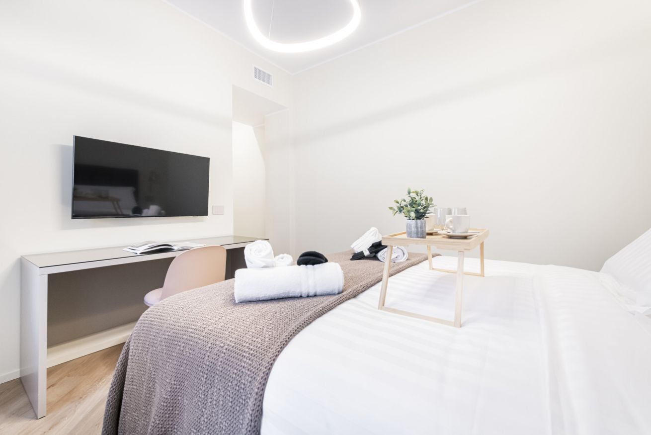 A modern hotel room with a bed, a desk, a flat-screen TV, and a bedside table with a plant and a cup.
