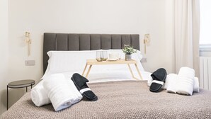 A neatly made bed with a grey headboard, a bedside table with a tray holding a glass, mug, and a small plant, and a pair of slippers and towels on the bed. - Easylife - Duomo Deluxe Apartments #1 (Milano)