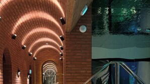 Area piscina interna moderna con parete curva in mattoni, zona di visuale in vetro e salottino con tavoli rotondi e illuminazione.