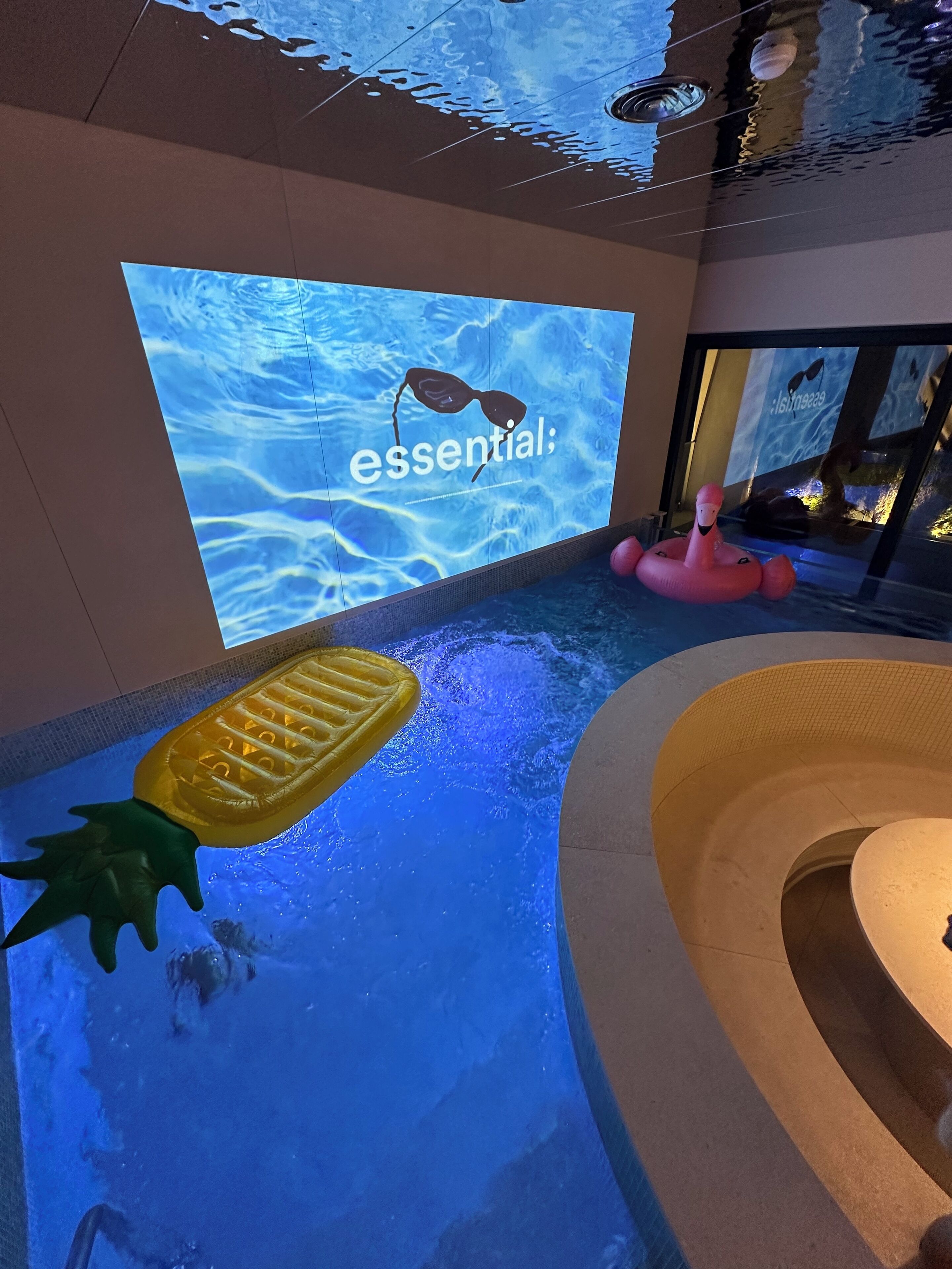 A pool area with a large screen displaying 'essential;' and a flamingo float.