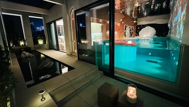 Area piscina interna moderna con pareti in vetro trasparente, una vasca idromassaggio e un piccolo angolo relax con una candela.